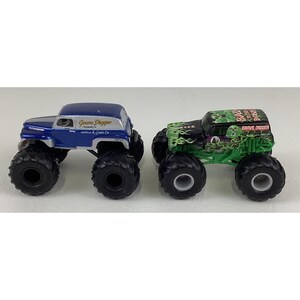 May include: Two toy monster trucks. One is blue and silver with "Grave Digger" text. The other is black and green with "Grave Digger" text and a skull graphic. Both have large black tires.