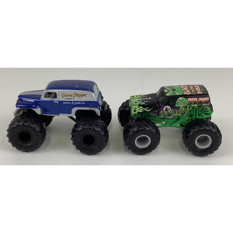 May include: Two toy monster trucks. One is blue and silver with "Grave Digger" text. The other is black and green with "Grave Digger" text and a skull graphic. Both have large black tires.