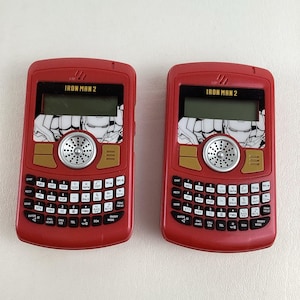 May include: Two red toy phones with a black and white image of Iron Man on the front. The phones have a keypad with numbers, letters, and symbols. The text "IRON MAN 2" is printed on the front of each phone.
