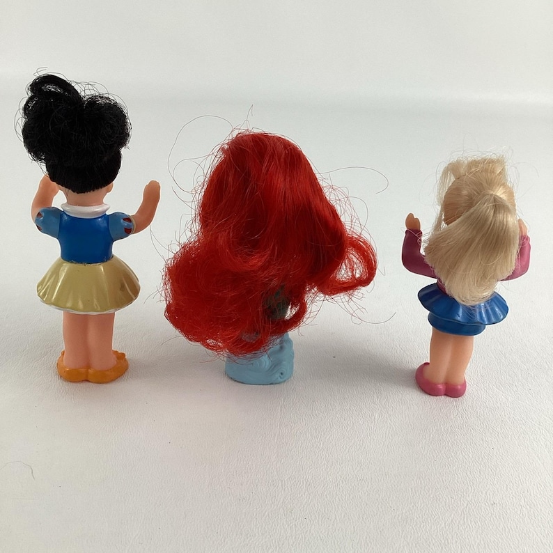 May include: Three small plastic dolls. One doll has black hair and wears a blue top and yellow skirt. The second doll has red hair. The third doll has blonde hair and wears a pink top and blue skirt.