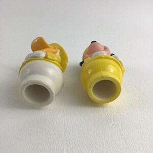 May include: Two plastic character figurines, one of Mickey Mouse and one of Donald Duck, both wearing yellow hats and sitting in yellow plastic tubs. The figurines are facing away from the camera, showing the bottom of the tubs.