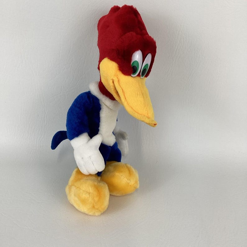Op de afbeelding: Een pluche Woody Woodpecker-speelgoed. Het speelgoed heeft een rode kop, een gele snavel en groene ogen. Het lichaam is blauw en wit, met gele voeten en witte handschoenen. Het speelgoed staat op een wit oppervlak.