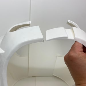 Op de afbeelding: Wit plastic pantserstuk met gebogen randen en een rechthoekig gedeelte. De afbeelding toont de montage van het pantser, met een hand die een losgemaakt gedeelte vasthoudt. De achtergrond is effen wit.