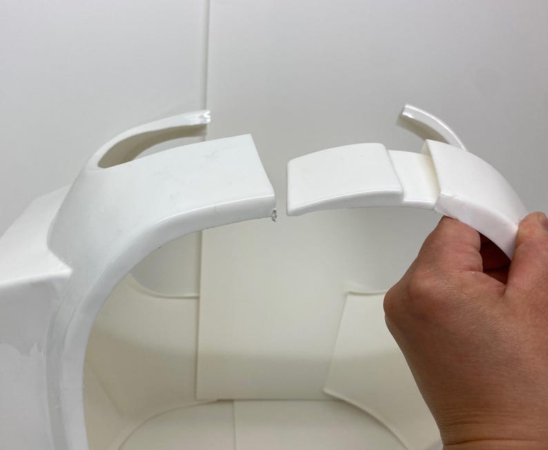 Op de afbeelding: Wit plastic pantserstuk met gebogen randen en een rechthoekig gedeelte. De afbeelding toont de montage van het pantser, met een hand die een losgemaakt gedeelte vasthoudt. De achtergrond is effen wit.