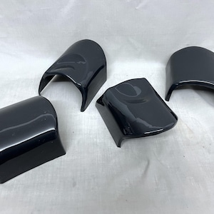 May include: Five glossy black automotive parts are arranged on a white surface. The parts have a curved, angular design, with a smooth, reflective finish. The shapes vary slightly, suggesting they are designed for different areas of a vehicle.