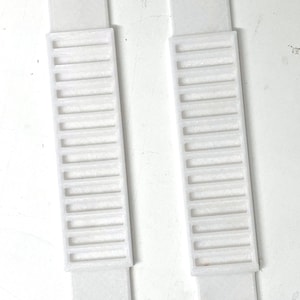 May include: Two white plastic strips with multiple rectangular holes. The strips are approximately 25 cm long and 2.5 cm wide.