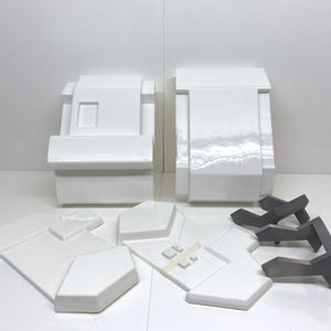 Può includere: Una collezione di componenti geometrici bianchi che include due grandi blocchi rettangolari, quattro pannelli angolari piatti con dettagli quadrati in rilievo e tre staffe metalliche grigio scuro a forma di Y disposte per l'assemblaggio.
