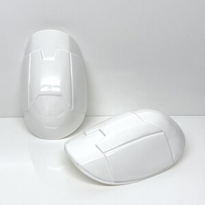 May include: Two white plastic knee pads with a textured surface and a raised design. The knee pads are designed for protection and support.