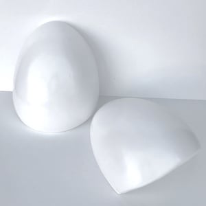 May include: Two glossy, white, egg-shaped decorative objects. One is upright, the other is tilted on its side. The objects have a smooth, reflective surface, and are set against a plain white background. The shapes are reminiscent of Easter or spring decor.