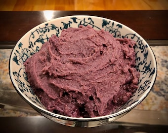 Può includere: Un primo piano di una ciotola di pasta di patate dolci viola. La pasta è liscia e cremosa, e la ciotola è decorata con un motivo floreale blu e bianco.