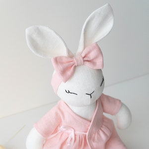 May include: A white plush bunny wearing a pink linen dress with a matching pink bow. The bunny has embroidered eyes and a stitched nose.