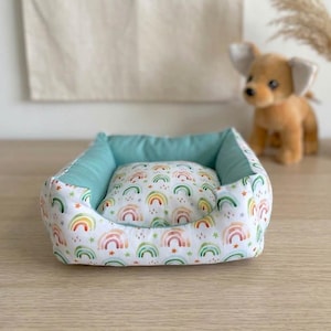 Puede incluir: Una pequeña cama cuadrada para mascotas con un colorido estampado de arcoíris sobre un fondo blanco. La cama tiene un interior azul claro y una almohada a juego. Un perro de juguete está en el fondo.