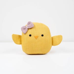 Op de afbeelding: Een gele pluche kuiken met een lavendelkleurige strik op het hoofd. Het kuiken heeft zwarte ogen en een kleine oranje snavel. De vleugels zijn uitgespreid, tegen een witte achtergrond.