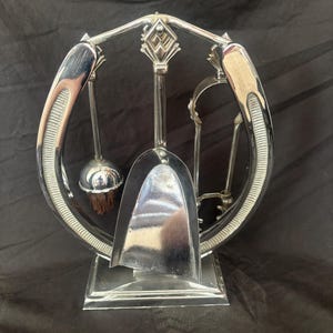 Peut inclure: Un ensemble d'outils de cheminée en chrome comprenant un support en forme de fer à cheval avec un fleuron décoratif central. Le set inclut une pelle, une brosse à poils sombres et une pince, suspendus dans le cadre sur une base rectangulaire.