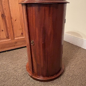May include: A cylindrical wooden cabinet in a deep reddish-brown hue. The cabinet features a rounded top and base, a single door with a brass-toned knob, and sits on a carpeted floor. This is a piece of furniture.