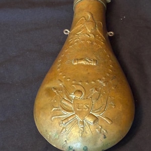 Könnte beinhalten: Antike Pulverflasche aus Messing mit geprägten patriotischen Motiven. Sie zeigt einen Adler, Sterne und ein Händedruck-Design. Die Flasche hat einen Ausguss und zwei kleine Schlaufen zum Aufhängen. Das US-Emblem ist auf der Vorderseite zu sehen.