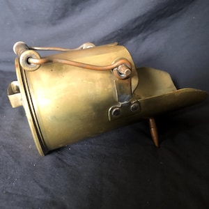 Puede incluir: Un objeto de estilo vintage en forma de cubo hecho de latón con un asa y una punta puntiaguda en la parte inferior. El objeto está hecho de latón y tiene un aspecto desgastado y antiguo.