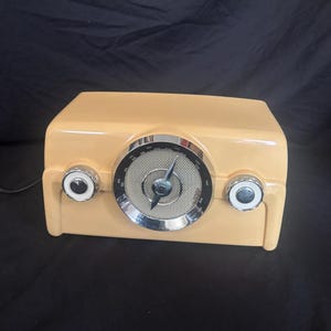 May include: A light peach-coloured tabletop radio with a prominent circular central speaker grille, a silver-toned dial with a black needle, and two smaller circular control knobs on the left and right sides, all encased in a smooth plastic housing.