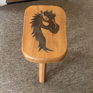 May include: A small wooden stool with a light brown finish and rounded corners. The top surface features a dark, detailed wood-burned illustration of a horse head with a flowing mane, centred on the seat. The stool has a single visible leg.