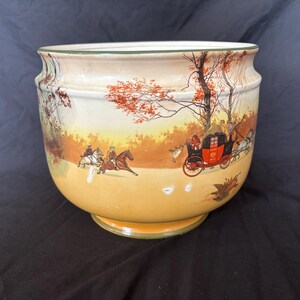 May include: A ceramic planter with a painted landscape scene of a horse-drawn carriage and riders. The planter has a wide, rounded shape with a pedestal base. The color palette includes cream, orange, and green.