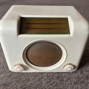 May include: Cream-coloured vintage radio with a gold mesh speaker cover and two white knobs. The radio has a rectangular display with a green and red scale. A black cord extends from the back.