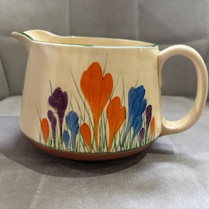 May include: A cream-coloured ceramic pitcher featuring a hand-painted floral design of orange, blue, and purple crocus flowers with green stems. The pitcher has a curved handle, a pouring spout, and a reddish-brown base rim.