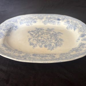 May include: An oval, antique serving platter with a white base and a blue floral pattern. The edges and center of the platter feature detailed floral designs. The platter is likely made of ceramic or porcelain.
