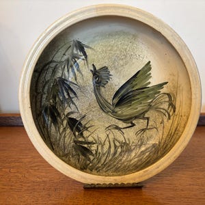 Puede incluir: Un plato redondo de cerámica con borde color crema y un centro beige texturizado y moteado. El plato presenta una ilustración pintada a mano de un ave crestada con plumaje verde oscuro y negro de pie entre hierbas altas y tallos de bambú.