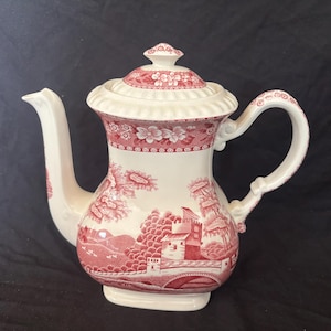 May include: A white and red ceramic teapot with a lid. The teapot features a scenic design with buildings, trees, and a bridge. The handle and spout are white. The lid has a floral pattern.