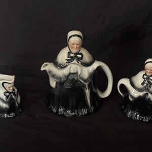 Puede incluir: Un juego de té de cerámica vintage que incluye una tetera, una jarra para la crema y un azucarero. El juego está pintado en blanco y negro, y cada pieza está adornada con la figura de una mujer con un gorro. La tetera tiene un asa y un pico curvos.