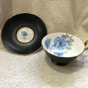 Könnte beinhalten: Ein Vintage-Tee-Set. Die Tasse und Untertasse sind schwarz mit goldenen Rändern. Die Innenseite der Tasse ist weiß mit blauen Blumenmustern. Die Untertasse hat ein passendes Blumenmuster in der Mitte.