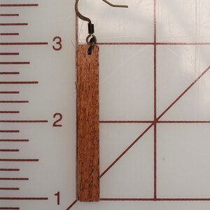 K&ouml;nnte beinhalten: Ein Holzohrring mit braunem Holzmaserungsmuster und einem goldfarbenen Haken. Der Ohrring ist etwa 5 cm lang.