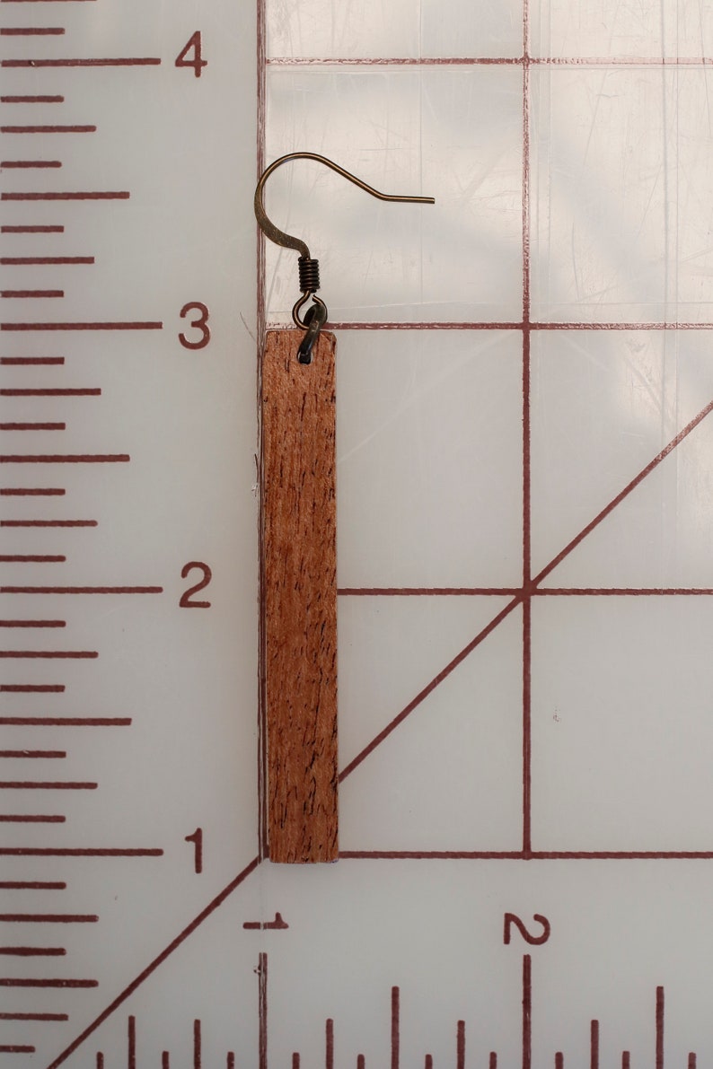 K&ouml;nnte beinhalten: Ein Holzohrring mit braunem Holzmaserungsmuster und einem goldfarbenen Haken. Der Ohrring ist etwa 5 cm lang.