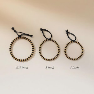 May include: Three beaded bracelets with black string ties. The bracelets are black and gold, with sizes of 16.5 cm, 12.7 cm, and 10.2 cm. The bracelets are displayed on a neutral background.