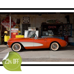 Puede incluir: Un Corvette convertible rojo y blanco de la década de 1950 estacionado frente a una gasolinera vintage de la Ruta 66. El coche tiene un techo blanco y molduras blancas. La gasolinera tiene un letrero que dice "Abierto" y "Ruta 66".