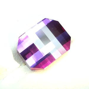 May include: A rectangular-shaped, faceted gemstone with beveled edges. The gem displays a vibrant colour palette of purple, magenta, and white, arranged in a geometric pattern. The gemstone is set against a white background.