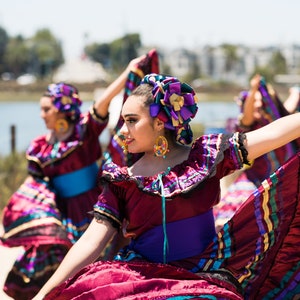 May include: Three women in colourful traditional Mexican dresses with purple, blue and gold accents. They are dancing and smiling, with their arms raised in the air.