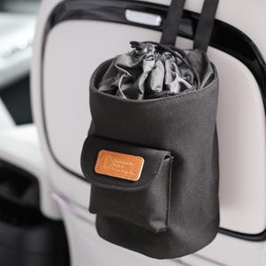 Könnte beinhalten: Schwarzer Autositz-Organizer mit Kordelzugverschluss und kleiner Fronttasche. Der Organizer wird an der Kopfstütze eines Autositzes aufgehängt. Die Vordertasche hat einen braunen Lederaufnäher mit einem Logo.