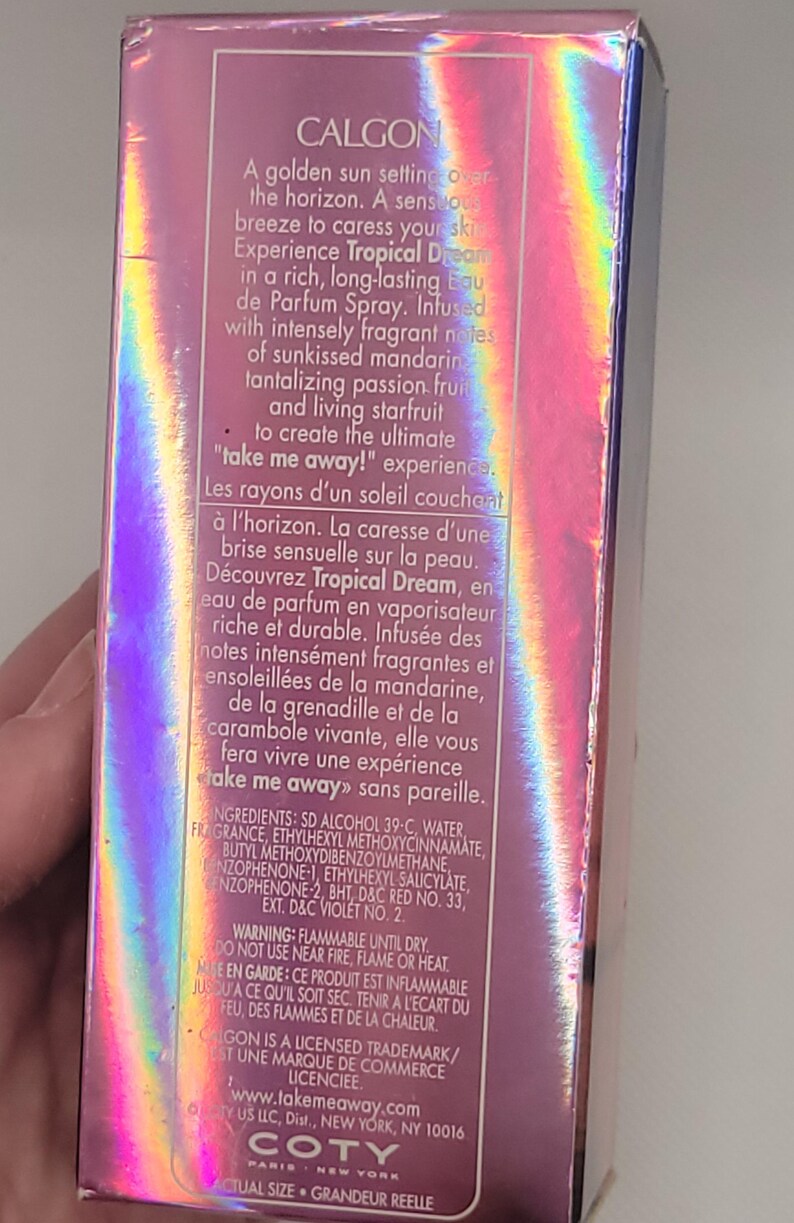 Puede incluir: Una caja rosa e iridiscente de Calgon Tropical Dream Eau de Parfum Spray. La caja presenta texto que describe las notas de mandarina, maracuy&aacute; y carambola. La marca y el logotipo son visibles.