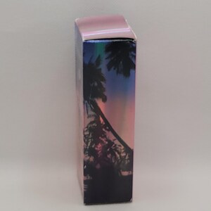Puede incluir: Una caja rectangular con una tapa iridiscente rosa y un dise&ntilde;o hologr&aacute;fico. El dise&ntilde;o presenta una silueta de palmeras contra un cielo de puesta de sol con tonos de rosa, morado y azul.