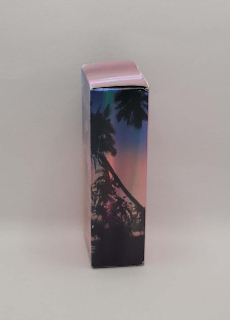 Puede incluir: Una caja rectangular con una tapa iridiscente rosa y un dise&ntilde;o hologr&aacute;fico. El dise&ntilde;o presenta una silueta de palmeras contra un cielo de puesta de sol con tonos de rosa, morado y azul.