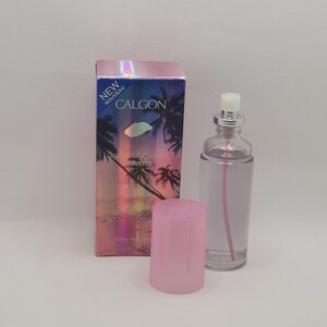 Puede incluir: Un frasco de perfume de vidrio transparente con tapa plateada y l&iacute;quido rosa, junto a una caja rosa. La caja tiene un dise&ntilde;o de puesta de sol tropical con palmeras y el texto "CALGON Tropical Dream Intense". El frasco tiene una tapa rosa.