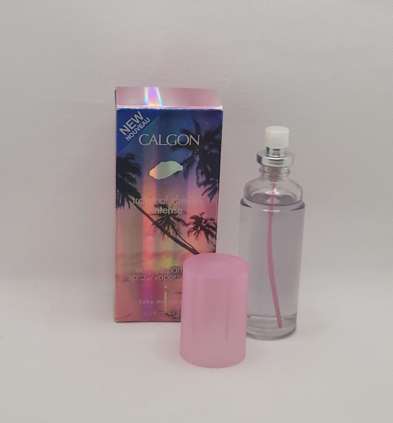Puede incluir: Un frasco de perfume de vidrio transparente con tapa plateada y l&iacute;quido rosa, junto a una caja rosa. La caja tiene un dise&ntilde;o de puesta de sol tropical con palmeras y el texto "CALGON Tropical Dream Intense". El frasco tiene una tapa rosa.