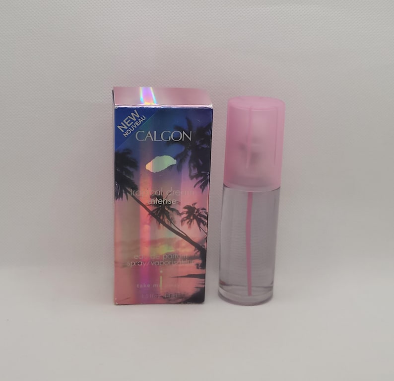 Puede incluir: Un frasco de perfume de vidrio transparente con una tapa rosa y una caja a juego. La caja presenta una escena tropical con palmeras y el texto "CALGON" y "NEW". El frasco de perfume es cil&iacute;ndrico y contiene un l&iacute;quido rosa.