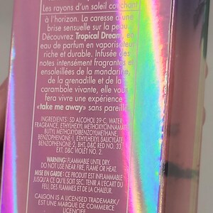 Puede incluir: Una caja de perfume con un acabado hologr&aacute;fico iridiscente. El texto en la caja incluye "Tropical Dream" y "take me away!" en ingl&eacute;s y franc&eacute;s. La caja es de color rosa y morado.
