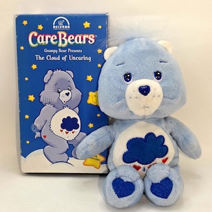 May include: A light blue Care Bear plush toy with a cloud belly and heart accents, next to a matching VHS tape. The tape box reads "Care Bears: Grumpy Bear Presents The Cloud of Uncaring" with star graphics.