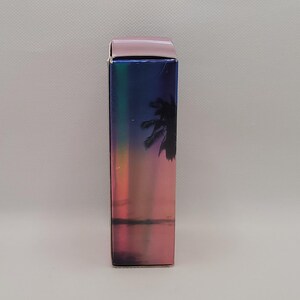 Puede incluir: Una caja rectangular con un acabado hologr&aacute;fico, que presenta una escena de puesta de sol con una silueta de palmera. La parte superior de la caja es de color rosa claro liso. La caja tiene una orientaci&oacute;n vertical.