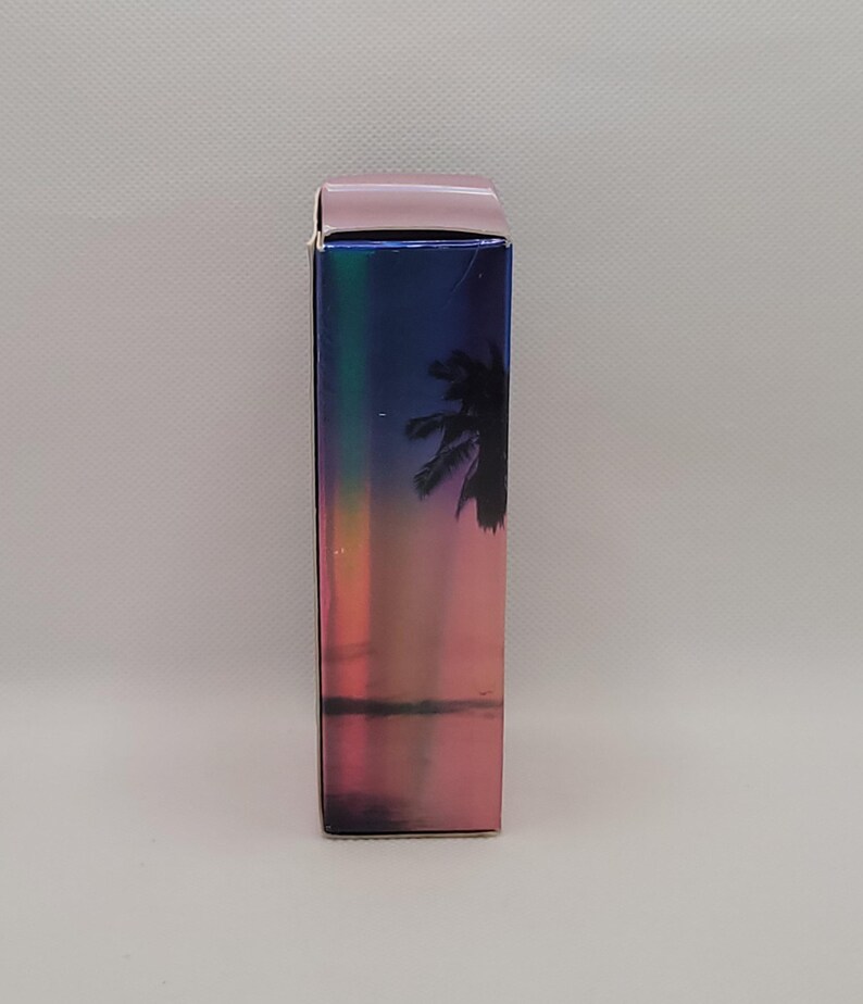 Puede incluir: Una caja rectangular con un acabado hologr&aacute;fico, que presenta una escena de puesta de sol con una silueta de palmera. La parte superior de la caja es de color rosa claro liso. La caja tiene una orientaci&oacute;n vertical.