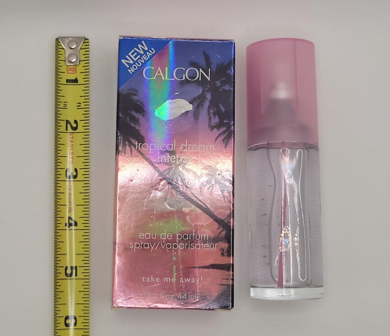 Puede incluir: Un frasco de spray de eau de parfum Calgon Tropical Dream Intense con tapa rosa y caja a juego. La caja presenta una escena tropical con palmeras y el texto "NEW NOUVEAU". El frasco es de 44 ml.