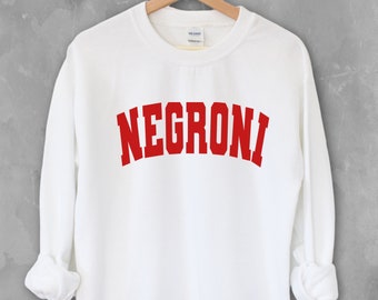 May include: White crewneck sweatshirt with the word "NEGRONI" printed in red, collegiate-style lettering. The long-sleeved top is hanging on a wooden hanger against a gray background.